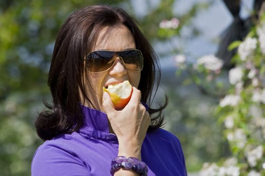 A mid-aged attractive woman is biting into a red-apple. In the background there are some blurred apple-trees. 