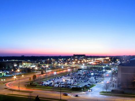 The streaming lights of cars as they enter and leave a busy shopping mall as the sun sets in the background.