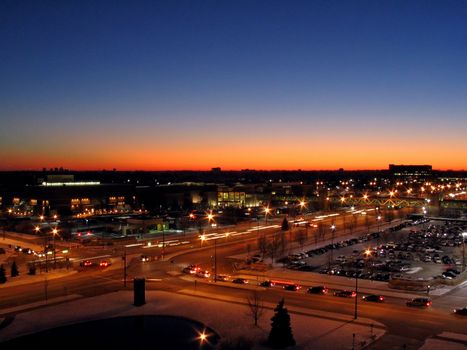 This is a night shot of a cross street in front of a shopping mall as the sun is setting in the background.