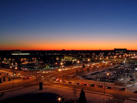 This is a night shot of a cross street in front of a shopping mall as the sun is setting in the background.