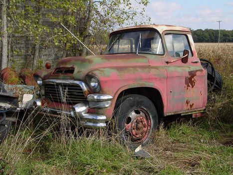 This is a picture of an old farm truck slowly decomposing back to its natural elements in a field, next to an old barn.