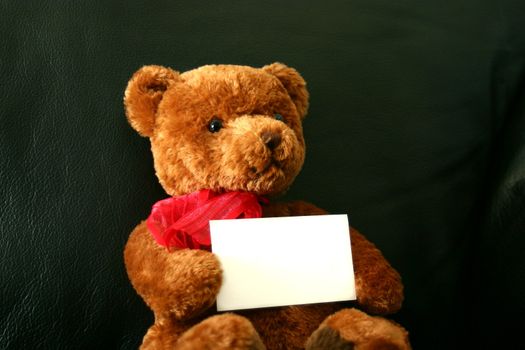 Soft brown teddy bear on a black leather couch with a red heart and red neckerchief holding a business card.