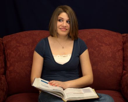 A pretty teenage girl sitting on a sofa studying a textbook.