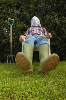 After a hard afternoon digging a gardener slumps back in a seat, exhausted, and covers his face with a hankerchief. Space for text on the background to the left, or on the grass of the foreground.