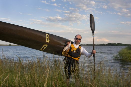 mature paddler carrying his long racing kayak on a grassy lake shore in northern Colorado, thirteen - temporary race number placed on deck by myself