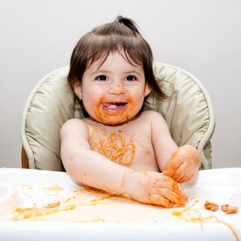 Happy baby having fun eating messy covered in Spaghetti Angel Hair Pasta red marinara tomato sauce.