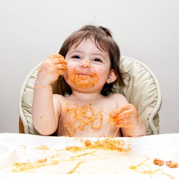 Happy smiling baby having fun eating messy covered in Spaghetti Angel Hair Pasta red marinara tomato sauce.