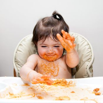 Happy baby having fun eating messy slapping hands covered in Spaghetti Angel Hair Pasta red marinara tomato sauce.
