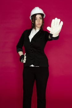 Young woman wearing white hard hat and construction glove, holding her hand up as a stop sign against a pink background