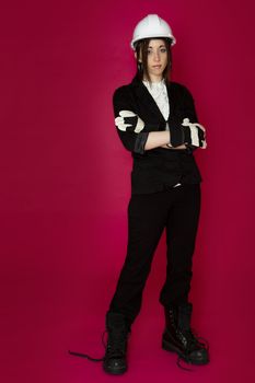 Young woman wearing white hard hat, construction glove and boots that are to big for her against a pink background
