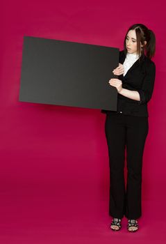 Young business woman wearing a business suit looking at a black board with surprise expression against pink background