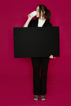 Young business woman wearing a business suit standing holding black board and drinking a coffee against pink background