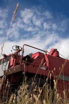 The front end of a red combine harvester machine stationary in a farm field in rural Wiltshire, England, set against a blue summer sky. Focus on grasses of field to front of image.