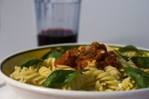 A plate full of italian pasta (fusilli al sugo), over a white background, with a glass of wine on the background