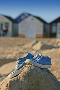 A pair of trainers/gym/beach shoes perched on top of some rocks at the beach, with beach huts in soft focus to the background.