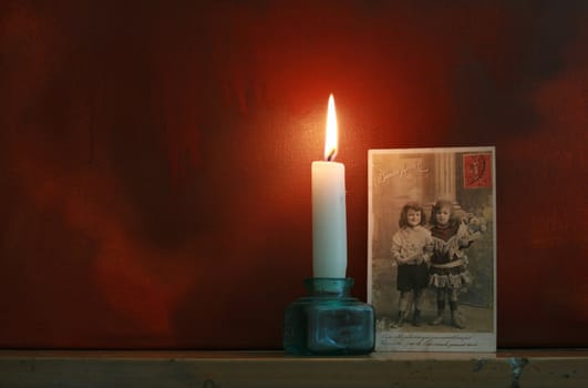 Victorian postcard set top a mantle piece with a candle against a dark red grunge styled background. Mood evocative.