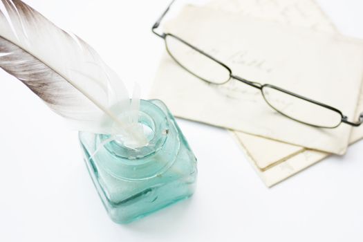 A feathered quill pen standing in a glass ink pot with old hand written letters behind with a pair of black framed reading glasses set on a white background