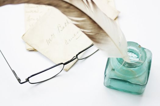 A feathered quill pen standing in a glass ink pot with old hand written letters behind, set on a white background