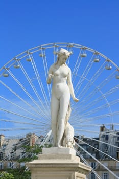 A statue in the Jardin Des Tuleres, Paris. Set against a 'ferris' type wheel to the background, against a clear blue sky, with green foliage and buildings.