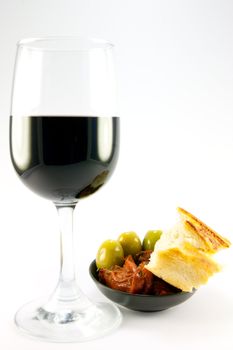 Pile of red sun dried tomatoes with three green olives and crusty bread in a small black dish with a glass of red wine on a plain background