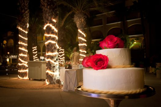 White wedding cake with red roses at night with lights wrapped around palm trees in the background.