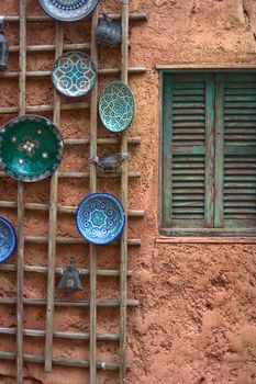 Painted blue and green plates hanging on a wodden frame attached to a exterior wall with a window that has wooden shutters on it