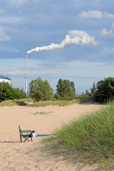 Chair standing on the beach and the industrial chimney with exhaust smoke in the background
