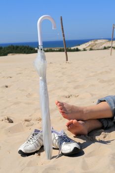 Woman's foot, pair of sneakers and umbrella on the sunny beach with the sea in background
