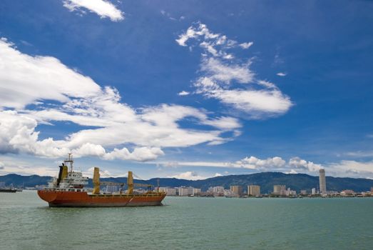 A large red cargo shipping vessel going through a strait and into the port of Penang in Malaysia. 
