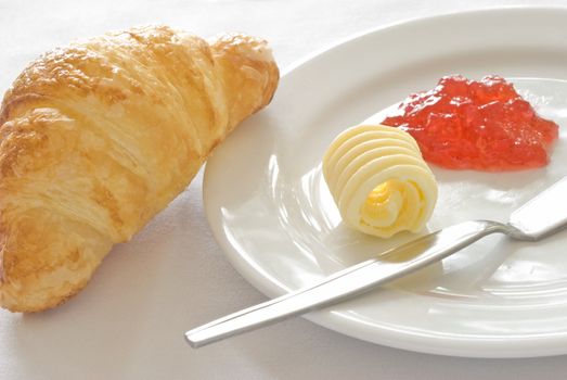 An image of a freshly baked croissant served on white ceramic plate with a portion of butter and jam. 