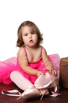 Little girl wearing a pink ballet outfit sitting next to an antique suitcase trying on oversized ballet shoes
