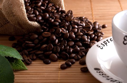 A bunch of coffee beans, falling out of a sack and a cup of fresh coffee on a wooden background