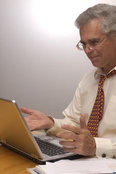 A businessman grins as he sits at his laptop computer, holding out his hands in satisfaction. he's done it!. Space for text on the blank wall behind him.
