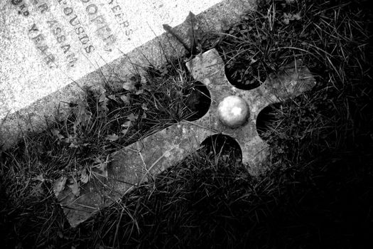 A black and white photograph of a cross, next to a tombstone.