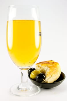 Glass of lager with three green olives and piece of crusty bread in a small black bowl on a plain background