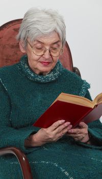 Portrait of an old woman reading a red book against a grey background.