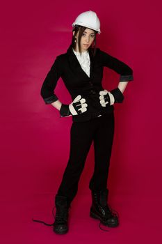 Young woman wearing white hard hat, construction glove and boots that are to big for her against a pink background