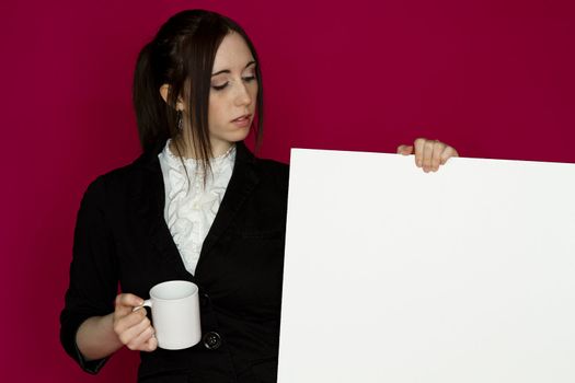 Young business woman holding a white board and a coffee cup with an inquiring expression against pink background