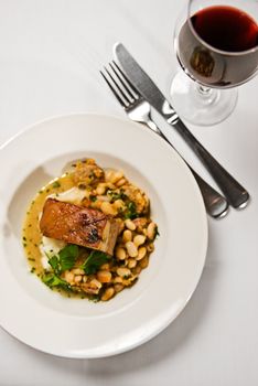 A peice of pork belly served with beans ang a glass of red wine. Restaurant food.