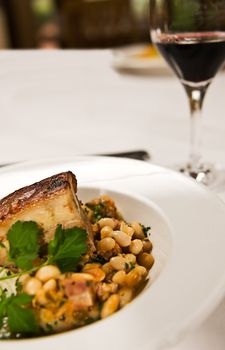 A peice of pork belly served with beans ang a glass of red wine. Restaurant food.