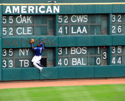 An outfielder for the Texas Rangers team attempts to catch a baseball by jumping at the wall. Ball is too high hitting just under the word American.