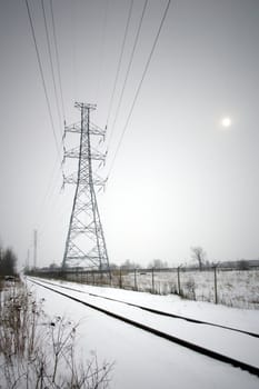 Focus on a large hydro tower which runs along railroad tracks in winter, snow covering the ground, with the sun visible in the haze.
