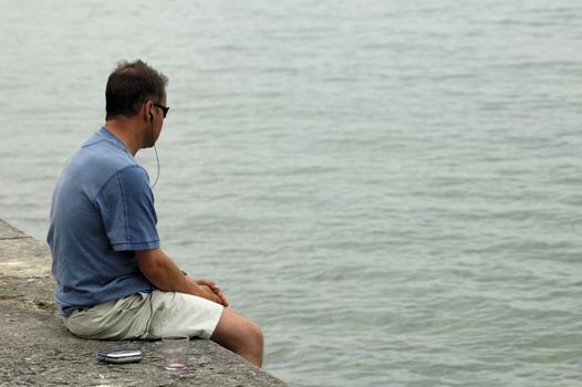 A solitary man, sitting on a sea wall with his hands clasped, gazing into the grey water thoughtfully. A music-player and empty wineglass are on the wall beside him.