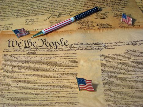 A copy of the Constitution of the United States resting on a copy of the Declaration of Independence accompanied by flags and a flag pen.