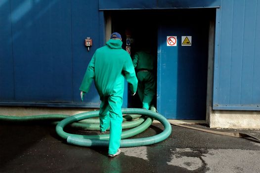 Two industrial workers wearing protection suits, entering the building. Vacuum tube on the ground.