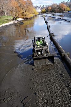 Worker in a small bulldozer excavating canal. Mud, pipeline, sky reflection in the water