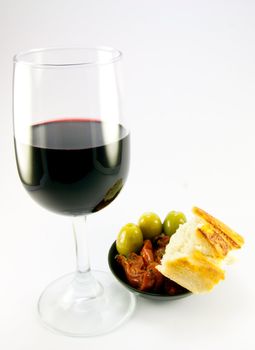 Pile of red sun dried tomatoes with three green olives and crusty bread in a small black dish with a glass of red wine on a plain background