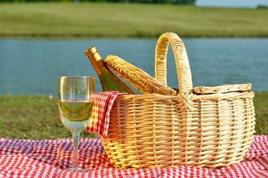 Bottle of white wine in picnic basket with glass of wine beside it.  Red gingham blanket and napkin with lake in background.