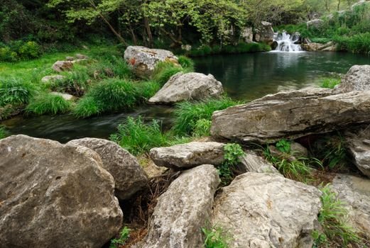 Image shows a small lake with a waterfall in the background and rocks in the foreground