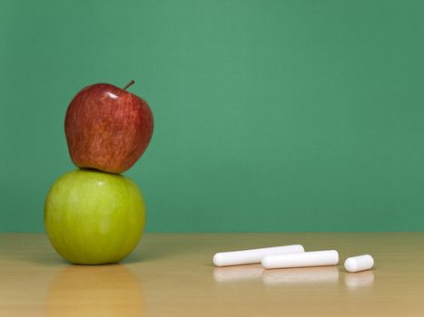 Some chalks and a red apple over a green one. A green chalkboard on the background.